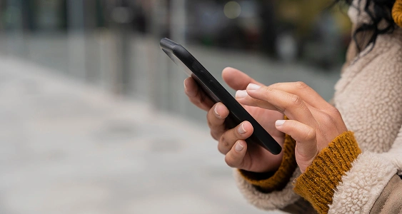 Close-up of hands with dark skin holding a black smartphone, touching the screen while standing outside in a cozy, sherpa-lined coat.